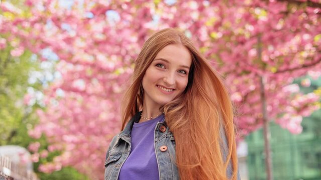 Portrait of young woman look around and smiling on blurred background in the city street. Sakura tree. Happy Caucasian female with long hair in casual denim jacket walking around Cherry blossom trees.