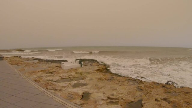 Public bench with statue of woman sitting by the sea in Torrevieja, Spain during a calima which is sand or dust suspended in the air. Gopro video of waves crashing against the rocks