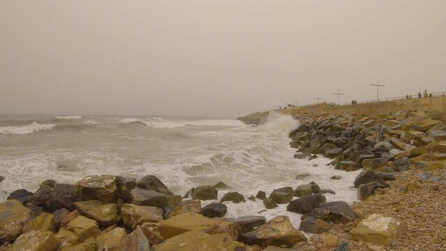 Gopro video of waves crashing against the rocks and heavy winds in Torrevieja, a tourist town at Costa Blanca Spain during a calima with people walking on a pier in the distance