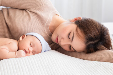 Obraz na płótnie Canvas Selective focus Asian mother lying on bed with newborn baby wearing blue hat in home, mom hugging little baby with love and care. Woman carying her adorable infant while toddler sleeping in bedroom.