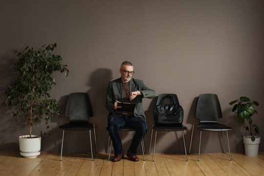 Mature man in eyeglasses checking time on his watch while sitting in waiting room and filling form