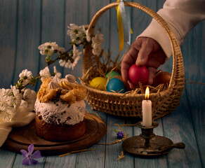 Easter cake, a basket with colored eggs, a bronze candlestick with a burning candle, a hand holding an egg and spring flowers with a blurred background