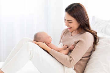 Beautiful Asian mother supports and tenderly cuddles the newborn baby gently while infant lying on lap. Mom looking and lifting baby with love and care. Toddler deeply sleeping on mother lap.