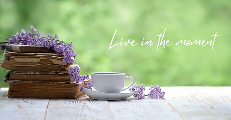 Live in the moment - inspiration quote. Cup of tea, flower and books on wooden white table, green natural background. spring season. tea party