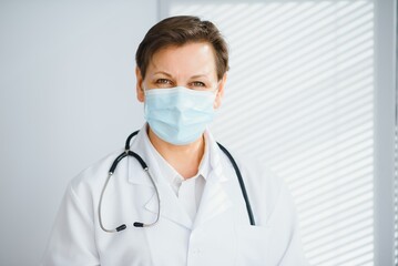 Older female doctor wearing face mask and white medical coat standing in hospital. Portrait