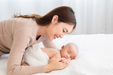 Fototapeta na wymiar Beautiful asian mom supports and tenderly cuddles newborn baby gently while baby deepy sleeping on bed. Young woman looking adorable infant with love and tender. Mother and child spend time together.