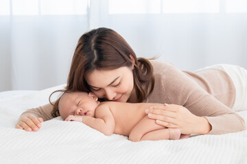 Obraz na płótnie Canvas Beautiful asian mom supports and tenderly cuddles newborn baby gently while baby sleep on bed. Young woman kissing babies with love and care. Mother and adorable infant spend time together at home.