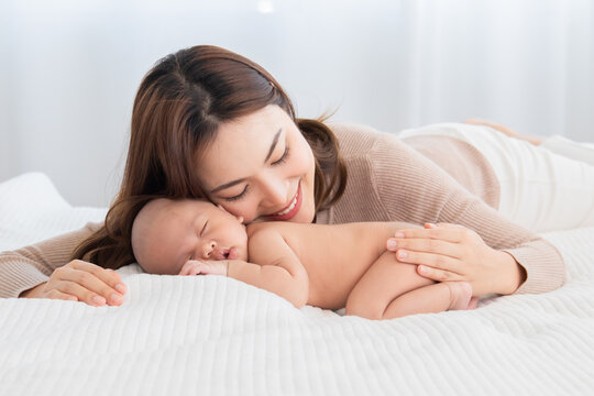 Beautiful Asian Mom Supports And Tenderly Cuddles Newborn Baby Gently While Baby Sleeping. Young Woman Kissing And Touching On Back Child With Love And Tender. Mother And Infant Spend Time Together.