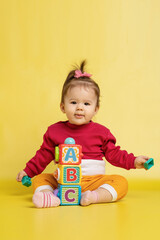 Girl 1 year old, playing with educational cubes with the English alphabet. The girl is sitting on the floor on a yellow background.