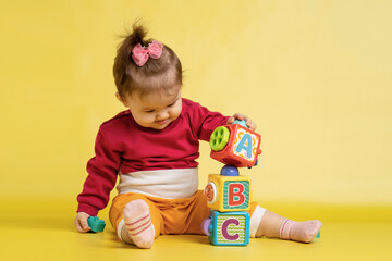 Girl 1 year old, playing with educational cubes with the English alphabet. The girl is sitting on the floor on a yellow background.