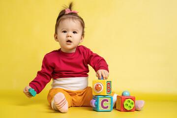 Girl 1 year old, playing with educational cubes with the English alphabet. The girl is sitting on the floor on a yellow background.