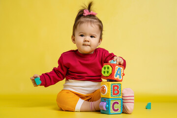 Girl 1 year old, playing with educational cubes with the English alphabet. The girl is sitting on the floor on a yellow background.