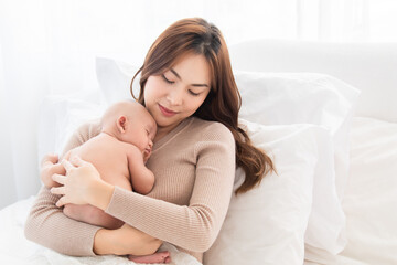 Beautiful Asian mother supports and tenderly cuddles newborn baby while toddler is sleeping on mom chest, Mother looking adorable infant with love and care. woman and child lying on bed together.