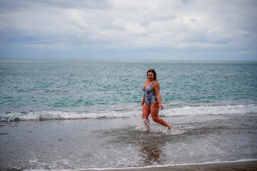 A plump woman in a bathing suit enters the water during the surf. Alone on the beach, Gray sky in the clouds, swimming in winter.