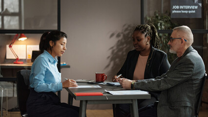 Young woman reading and signing a contract for applying a job while sitting at table with employers