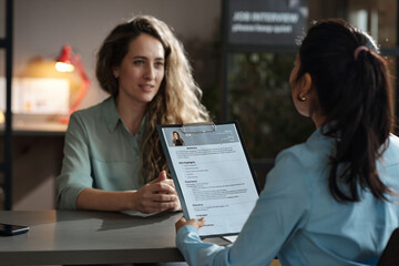 Rear view of businesswoman examining resume of young girl while she sitting at table and telling about herself at interview