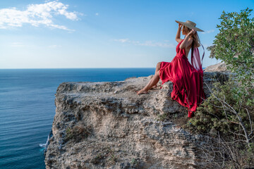 A girl with flowing hair in a long red dress sits on a rock above the sea. The stone can be seen in the sea.