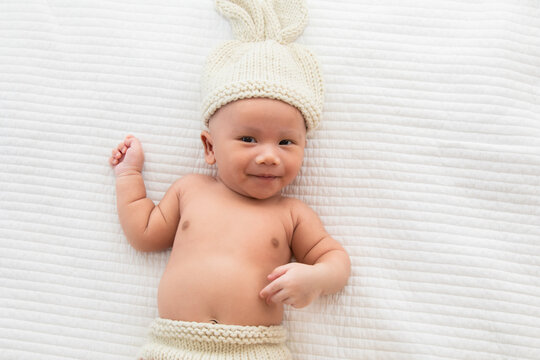 Asian adorable newborn baby wearing pants and rabbit hat beige knitted lying on white bed. little baby 0-1month play and talk with camera. Baby products concept. Happy childhood and motherhood.