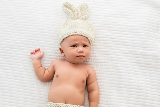 Asian adorable newborn baby wearing pants and rabbit hat beige knitted lying on white bed. little baby 0-1month looking surprised at camera. concept of happy childhood and motherhood.