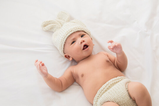 Asian adorable newborn baby wearing pants and rabbit hat beige knitted lying on white bed. little baby 0-1month play and talk with camera. Baby products concept. Happy childhood and motherhood.