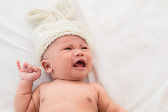 Asian Newborn wearing pants and rabbit hat beige knitted tired and hungry in bed. Healthy little kid shortly after birth. Adorable infant 0-1month crying sick fever lying on white bed.