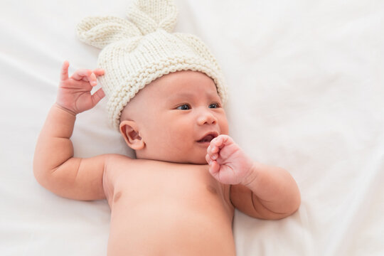 Asian Newborn wearing pants and rabbit hat beige knitted tired and hungry in bed. Healthy little kid shortly after birth. Adorable infant 0-1month crying sick fever lying on white bed.