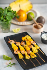Grilled shrimp skewers with pineapple and mango. Shrimp skewers with mint, pineapple, mango, lime and yogurt sauce on a black plate.