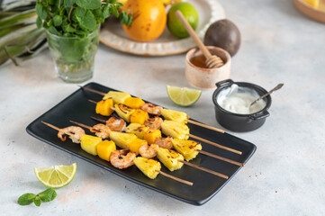 Grilled shrimp skewers with pineapple and mango. Shrimp skewers with mint, pineapple, mango, lime and yogurt sauce on a black plate.