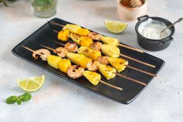Grilled shrimp skewers with pineapple and mango. Shrimp skewers with mint, pineapple, mango, lime and yogurt sauce on a black plate.