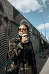 portrait of attractive young woman in trendy clothes and in sunglasses with paper cup coffee cup in hand. Female model posing on the painted wall background