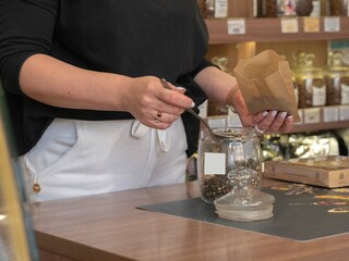 The seller of the store girl pours fragrant tea from different herbs with a spoon from a glass jar with a paper bag. Business concept for retail sales of tea and coffee