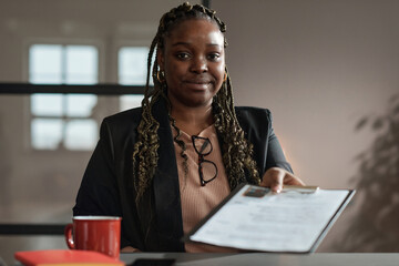 Portrait of African young woman with braids looking at camera while sitting at table and giving her...