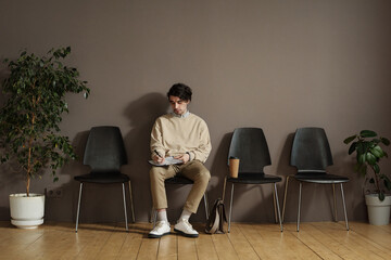 Young man sitting on chair in waiting room and filling form before job interview