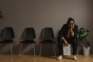 African young woman sitting with resume in waiting room alone and waiting for her turn in job...