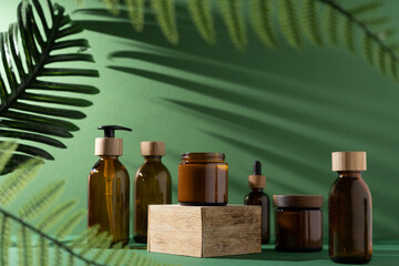 Obraz na płótnie Canvas Bottles for cosmetics and cream on a pedestal, green background with leaves and shadow. Brown glass jars with wooden lids. Eco concept. Container for shampoo, oil. Bottle with dispenser and pipette.