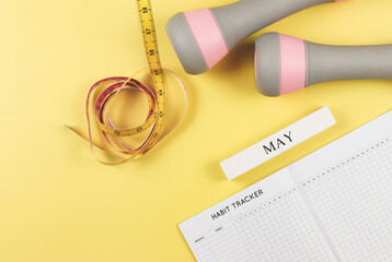 flat lay of habit tracker book, wooden calendar May,  gray pink dumbbells and measuring tape  on...