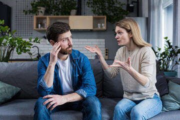 Family quarrel, woman quarrels and yells at the loser's husband, couple at home sitting in the living room on the couch - obrazy, fototapety, plakaty