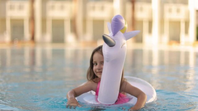 Child in swimming pool on unicorn inflatable violet ring. Little girl learning to swim with float. Water toy for baby and toddler. Healthy outdoor sport activity for children. Kids beach fun.