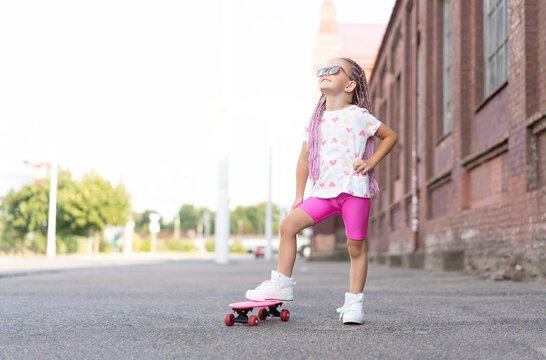 Fashion child girl in sunglasses with pink skateboard in the city.