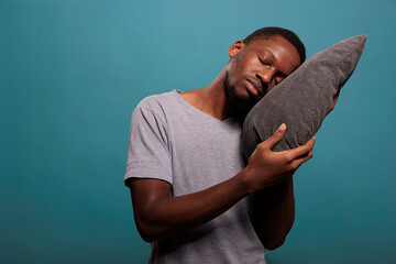 Tired man using comfortable pillow to get sleepy and take nap over blue background. Exhausted...
