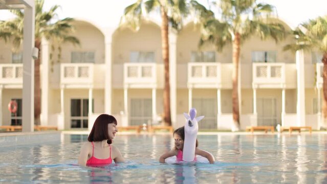 Little girl with unicorn inflatable ring with her mom in the same pink swimsuits relaxing in a swiming pool at sunny day. Happy mother and daughter in the pool at the leisure center. Copy space.
