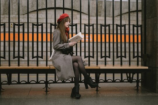 Beautiful Caucasian woman wearing coat, beret and scarf sitting alone on Saint Petersburg metro station reding a book. Image with selective focus and noise effect