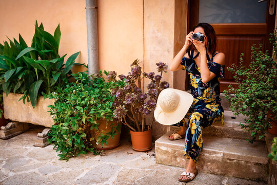young woman with red hair sitting on a door step with a camera in from of her face and taking a photo, next to potted plants