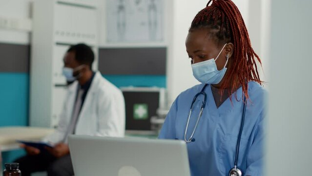 Portrait Of African American Nurse With Face Mask Working On Laptop, Using Technology To Plan Patient Appointments. Medical Assistant Sitting At Cabinet Desk For Health Care Service.