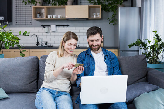 Happy married couple man and woman sitting at home on the couch using laptop for online shopping, holding a credit bank card