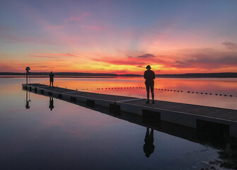 Fototapeta premium person on the pier at the calm lake, summer time, beautiful orange and pink sunset. Tranquil holidays concept.