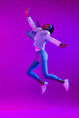 Fototapeta na wymiar Full body portrait of ecstatic African-American woman jumping in modern neon light studio background