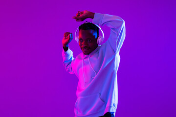 Young African man wearing wireless headphones listening to music and dancing in studio modern neon...
