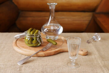 A glass of vodka is on the table next to pickles and a decanter on the background of a log wall. 