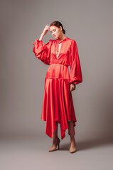 Bright young lady in red oversize dress with long wide sleeves and sexy neckline on high heels mooving on grey studio background. Elegant contemporary bohemian female look.